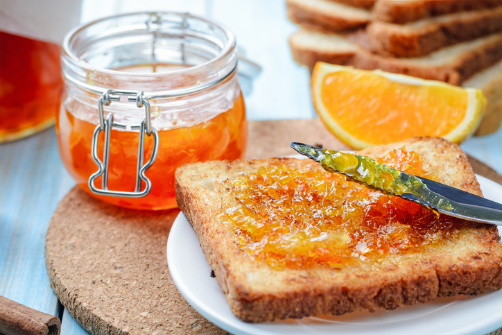 Ein Glas Orangenmarmelade neben einer Scheibe Toast, auf die Marmelade mit einem Messer gestrichen wird.