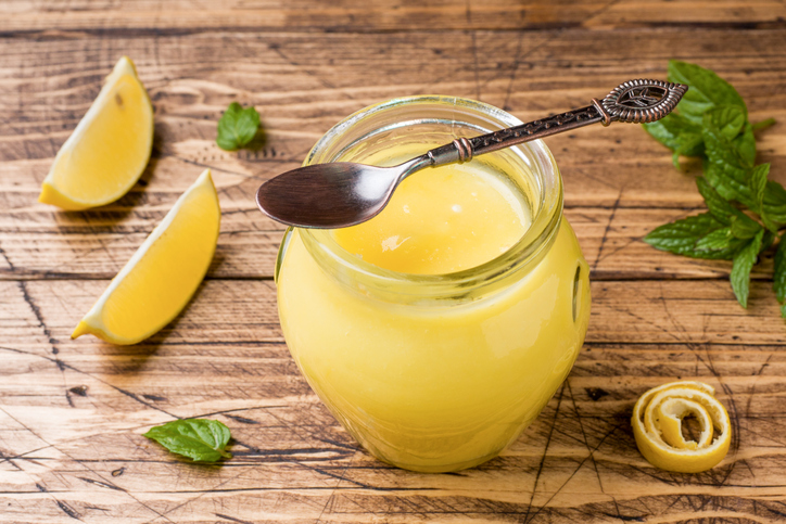 Draufsicht auf ein Glas Zitronencreme mit einem Löffel, garniert mit Minzblättern und einer kunstvoll gedrehten Zitronenzeste.
