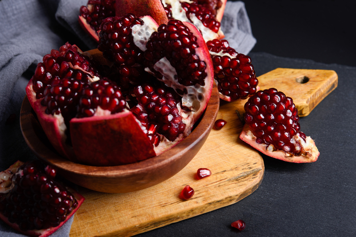 Mehrere Stücke von Granatapfel mit dunkelroten Kernen liegen in einer Holzschale und auf einem Brett.
