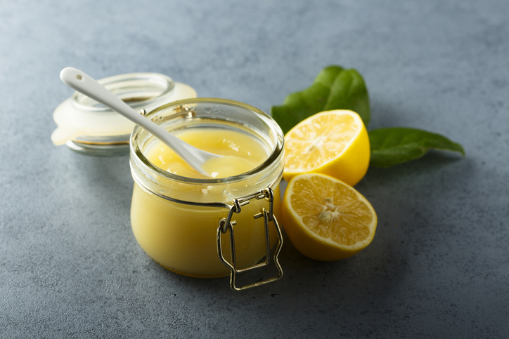 Ein offenes Glas Zitronencreme auf einer grauen Arbeitsfläche mit angeschnittenen Zitronen im Hintergrund.