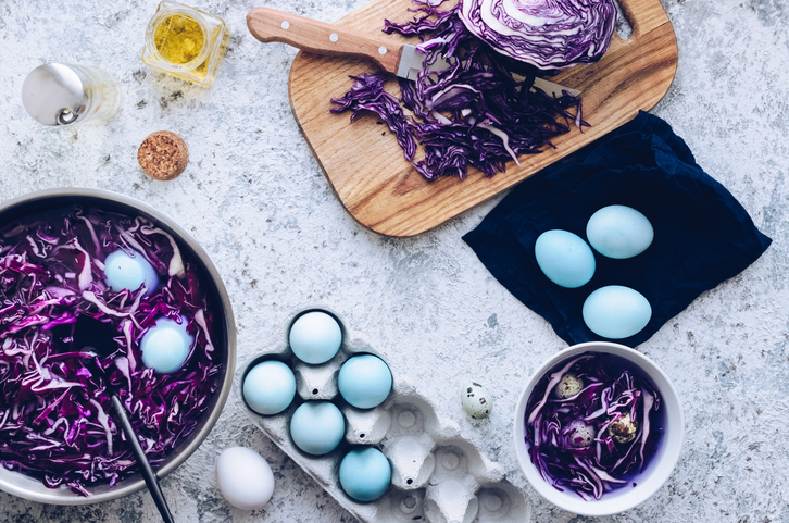 Eine Draufsicht zeigt blau gefärbte Eier in einem Eierkarton und in einem Topf mit violettem Rotkohlsud auf einem hellen Untergrund.