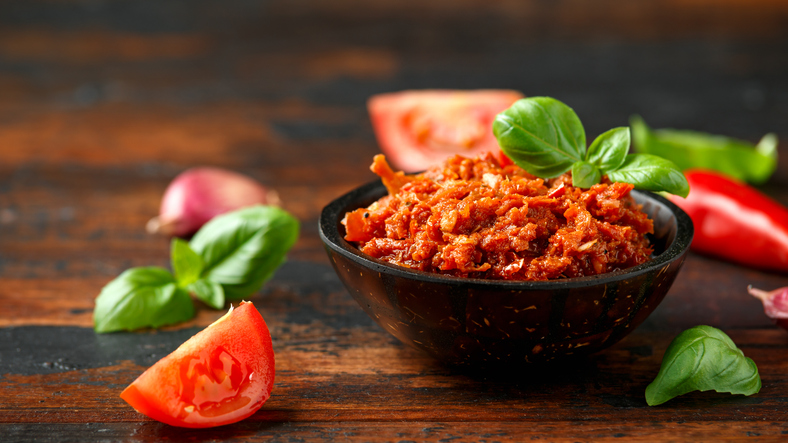 Eine kleine dunkle Schale mit Pesto Rosso auf einer dunklen Holzoberfläche, umgeben von Knoblauch, Tomaten und Basilikum.