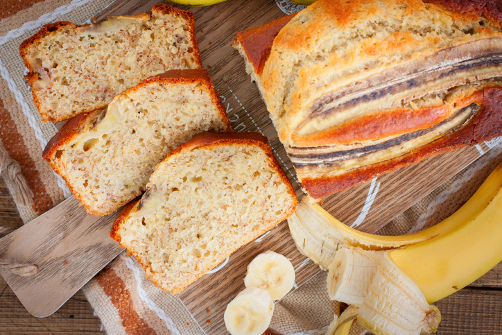 Mehrere Scheiben Bananenbrot liegen neben einem ganzen Laib und einer geschälten Banane auf einem Holzbrett.