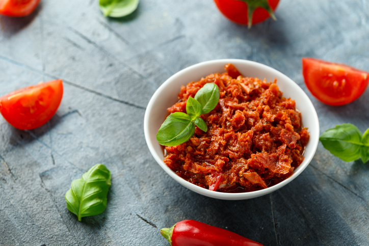 Draufsicht auf eine weiße Schüssel mit Tomatenpesto und Basilikumblättern auf einem grauen Hintergrund mit Tomatenstücken.