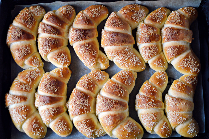 Draufsicht auf ein Backblech mit vielen gleichmäßig angeordneten Sesamcroissants auf Backpapier.