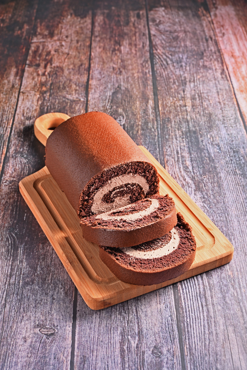 Eine Schokoladen-Biskuitrolle mit Cremefüllung liegt auf einem Holzbrett, daneben sind zwei Scheiben abgeschnitten.