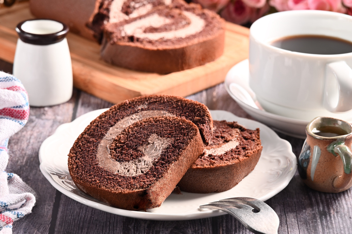 Eine Scheibe Schokoladen-Biskuitrolle mit Creme liegt auf einem weißen Teller neben einer Tasse Kaffee.