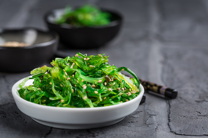 Hellgrüner Wakame-Algensalat mit Sesamkörnern in einer weißen Keramikschale auf dunklem Stein.