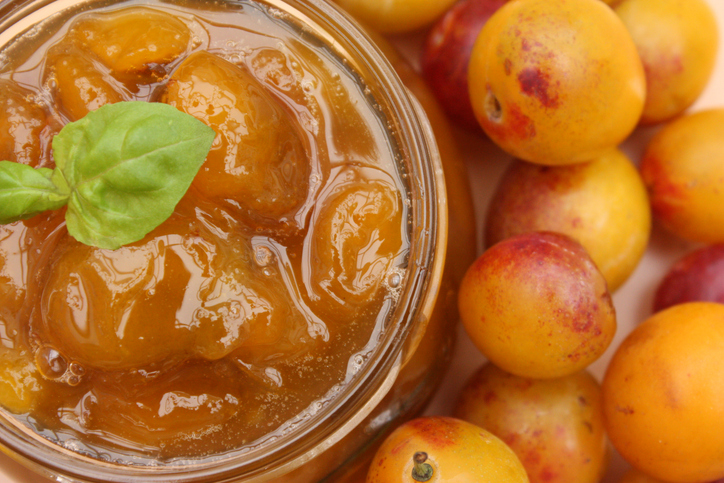 Ein Glas mit goldgelber Mirabellenmarmelade, garniert mit einem Minzblatt, neben frischen, reifen Früchten.