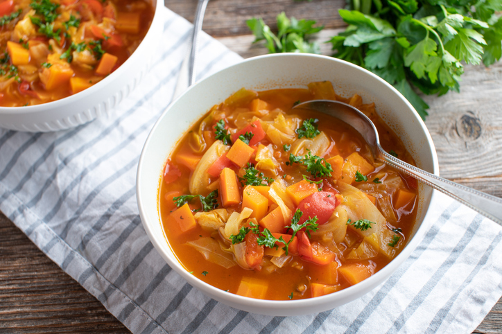 Eine Schale Kohl-Gemüsesuppe mit einem silbernen Löffel auf einer blau-weiß gestreiften Serviette vor frischer Blattpetersilie.