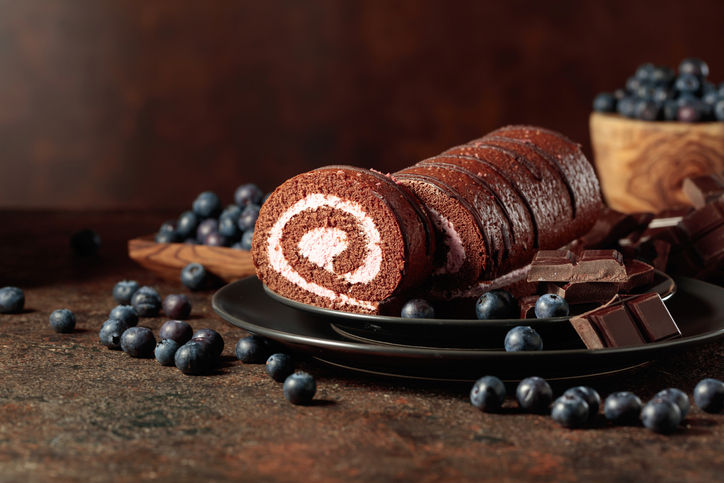 Eine Schokoladen-Biskuitrolle mit heller Creme liegt auf einem Teller zwischen Heidelbeeren und Schokoladenstücken.