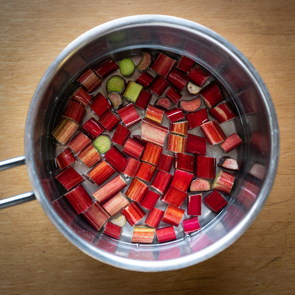 Draufsicht auf einen Edelstahltopf mit Wasser und gleichmäßig geschnittenen, tiefroten Rhabarberstücken.