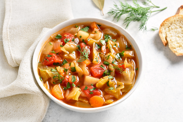Eine weiße Schale Gemüsesuppe auf einer hellen Tischdecke neben knusprigen Baguettescheiben und frischem Dill.