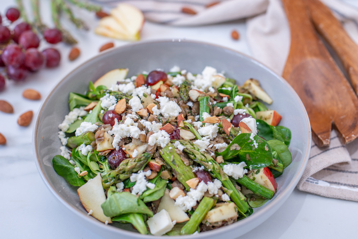 Eine Schüssel mit Grünem Spargelsalat, weißen Käsekrümeln, roten Trauben, Apfelstücken und gehackten Mandeln steht auf einem Tisch.
