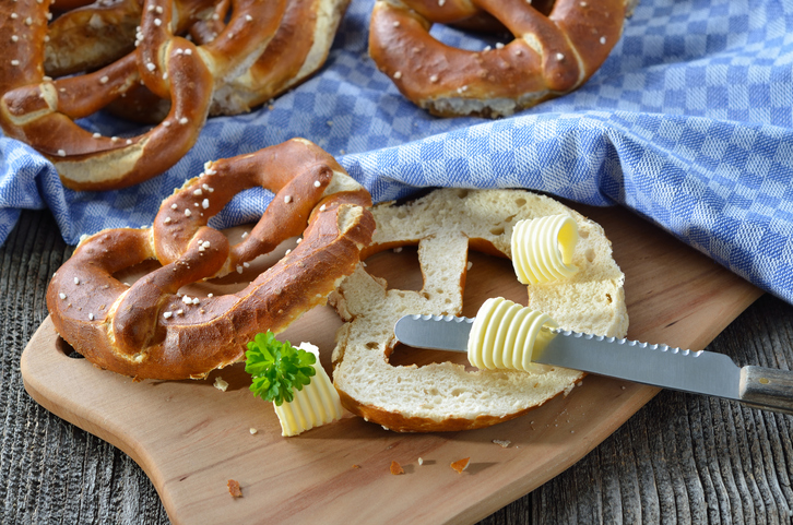 Eine aufgeschnittene Brezel liegt mit Butter und Messer auf einem Holzbrett, weitere Brezeln sind im Hintergrund zu sehen.