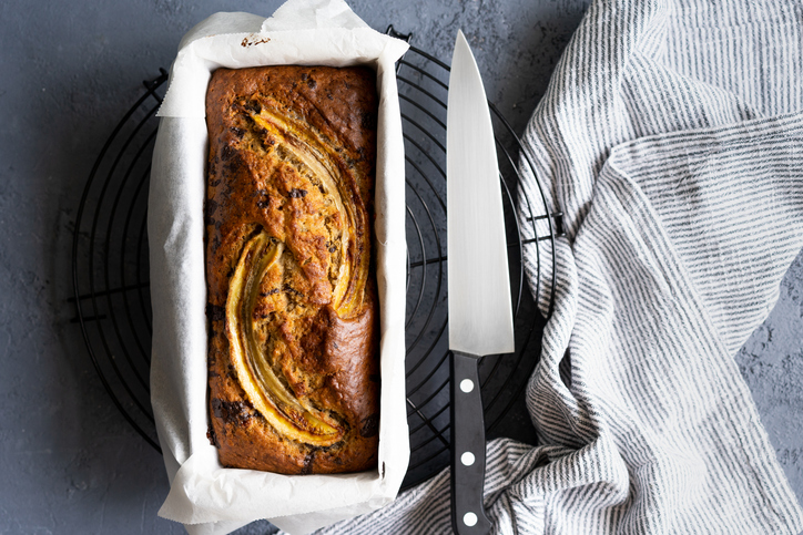 Von oben ist ein Bananenbrot in der Form neben einem Messer und einem Küchentuch zu sehen.