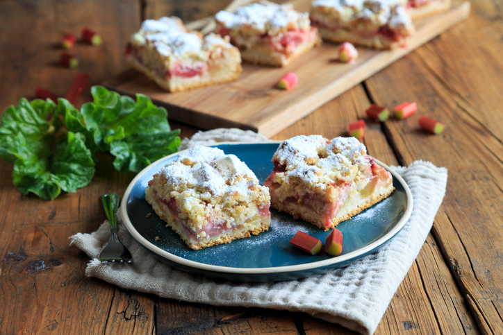 Eine Servierszene mit Rhabarberkuchen-Stücken auf einem Holzbrett und einem blauen Teller, dekoriert mit frischen Rhabarberblättern.