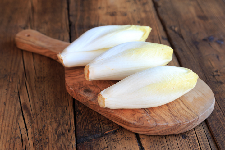 Drei makellose, weiße Chicorée-Köpfe mit gelben Spitzen auf einem rustikalen Holzschneidebrett.