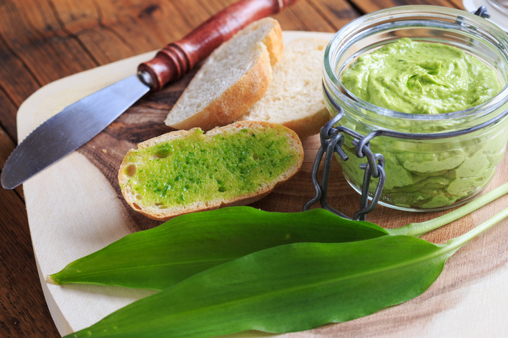Ein Glas mit cremiger Bärlauchbutter steht neben einer bestrichenen Brotscheibe und frischem Bärlauch.