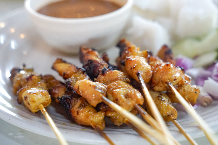 Mehrere Chicken Satay Spieße liegen auf einem Teller neben einer hellbraunen Sauce.