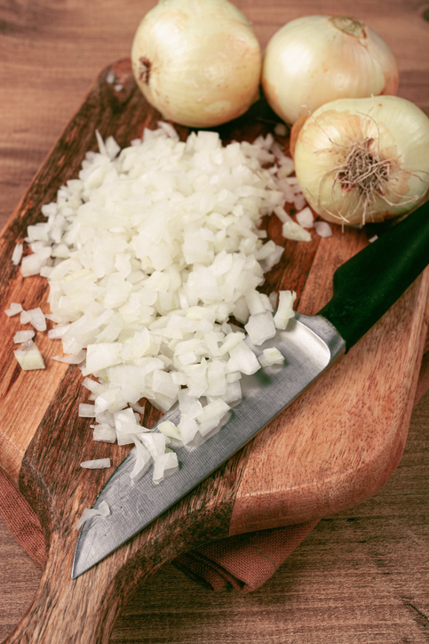 Gehackte Zwiebel liegt auf einem Holzbrett neben ganzen Zwiebeln und einem Messer.