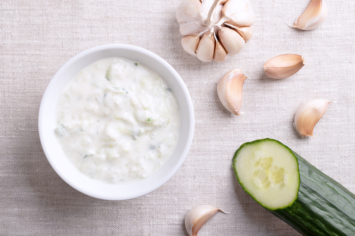 Draufsicht auf eine weiße Schale Tzatziki, umgeben von einer ganzen Knoblauchknolle, einzelnen Knoblauchzehen und einem Stück Salatgurke.