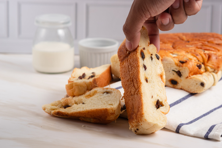 Eine Hand hält eine Scheibe Rosinenbrot vor einem angeschnittenen Laib auf dem Tisch.