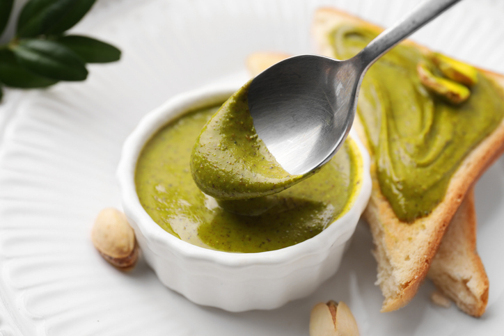 Ein Löffel hebt Pistaziencreme aus einer kleinen weißen Schale, daneben liegt Toastbrot mit grünem Aufstrich.
