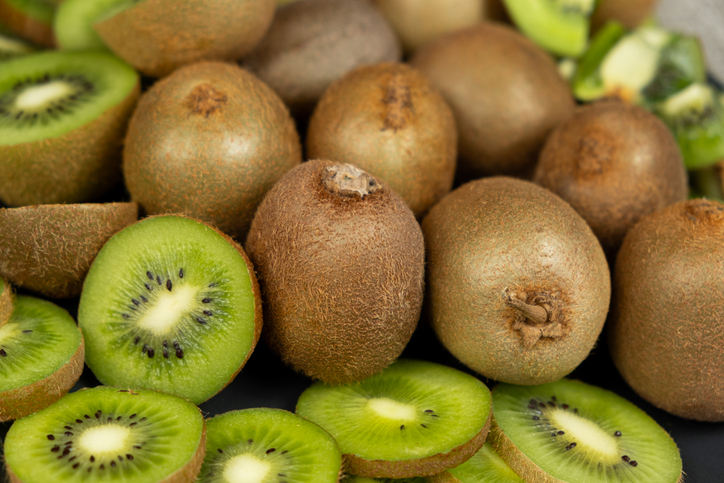 Mehrere braune Kiwis liegen zusammen mit vielen aufgeschnittenen grünen Kiwi-Stücken auf dunklem Untergrund.