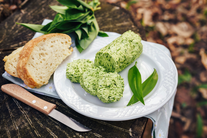 Auf einem weißen Teller liegen Bärlauchbutter in Scheiben, Brot und frische Bärlauchblätter.