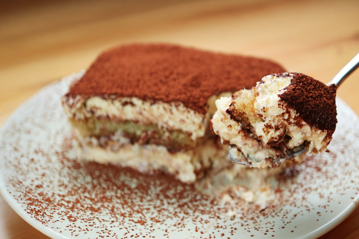 Ein Löffel hebt ein Stück luftiges Tiramisu mit sichtbaren Schichten aus Mascarponecreme und Kaffee-Biskuits an.