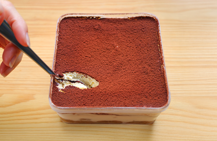 Eine Hand sticht mit einem Löffel in eine rechteckige Form mit Tiramisu, das eine gleichmäßige Kakaoschicht trägt.