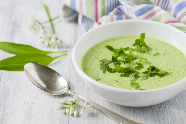 Eine Schale mit Bärlauchsuppe steht neben einem Löffel und frischem Bärlauch auf hellem Untergrund.
