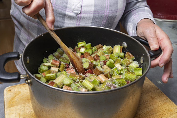 Frische Rhabarberstücke vermengt mit Zucker in einem großen Kochtopf, die mit einem Holzlöffel umgerührt werden.