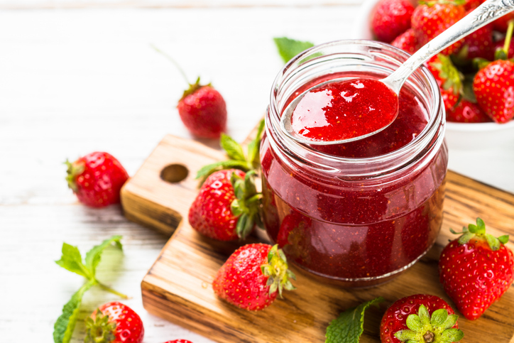 Ein Glas Erdbeermarmelade mit Löffel steht auf einem Holzbrett neben frischen Erdbeeren.