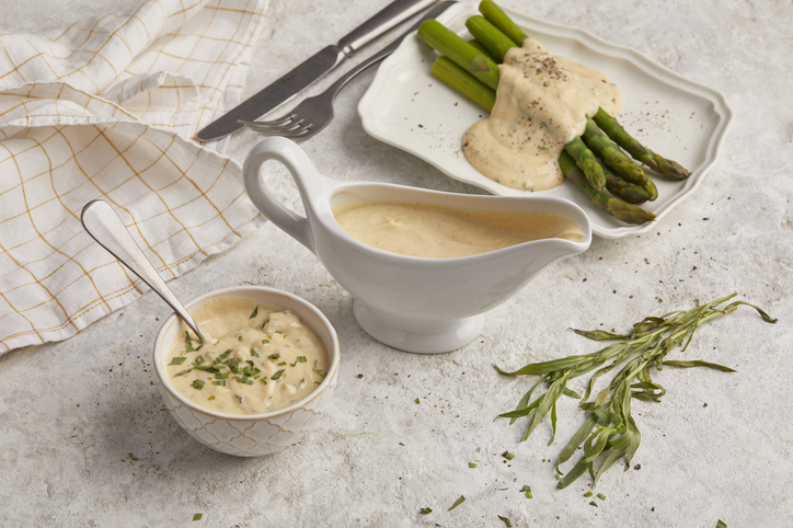 Eine Sauciere und ein Teller mit Spargel und Sauce Hollandaise stehen auf einem hellen Tisch.
