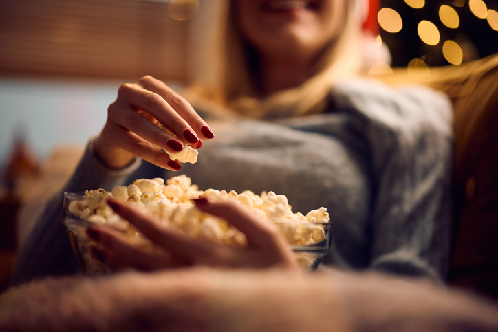 Eine Person greift mit der Hand in eine Glasschüssel voller Popcorn auf dem Sofa.