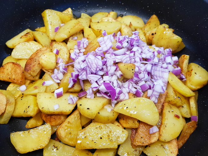 Gebratene Kartoffeln in einer Pfanne, über die eine Handvoll frisch gewürfelte rote Zwiebeln gestreut wurde.