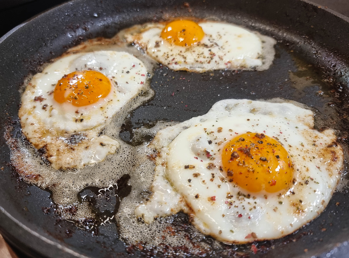 Drei frisch in der Pfanne bratende Spiegeleier, großzügig mit buntem Pfeffer und Gewürzen bestreut.