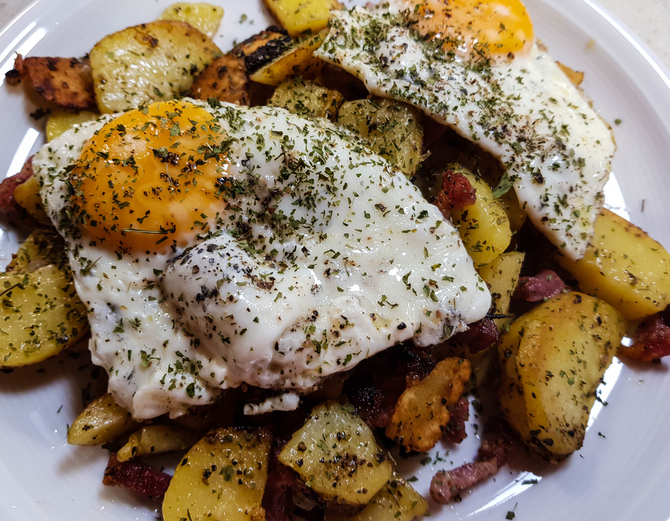 Ein Teller mit knusprigen Bratkartoffeln, Speck und zwei darauf platzierten Spiegeleiern, garniert mit getrockneten Kräutern.