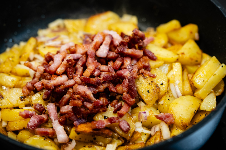 Nahaufnahme von in der Pfanne gebratenen Kartoffelspalten, die mit einer ordentlichen Portion gewürfeltem Speck getoppt sind.