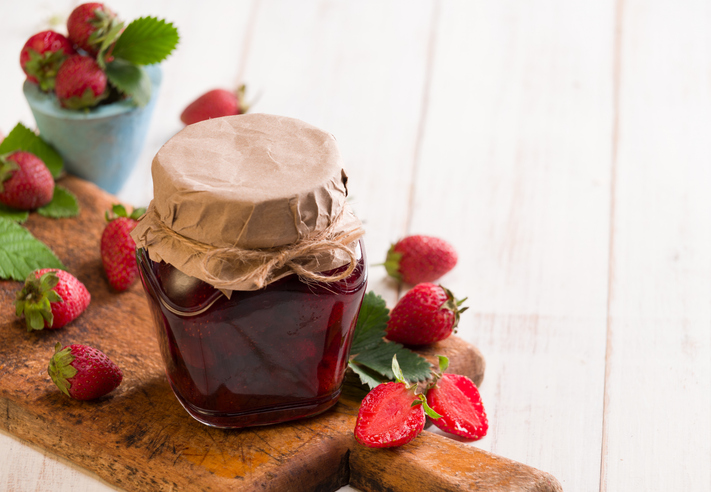 Ein verschlossenes Glas Erdbeermarmelade steht auf einem Holzbrett neben frischen Erdbeeren.