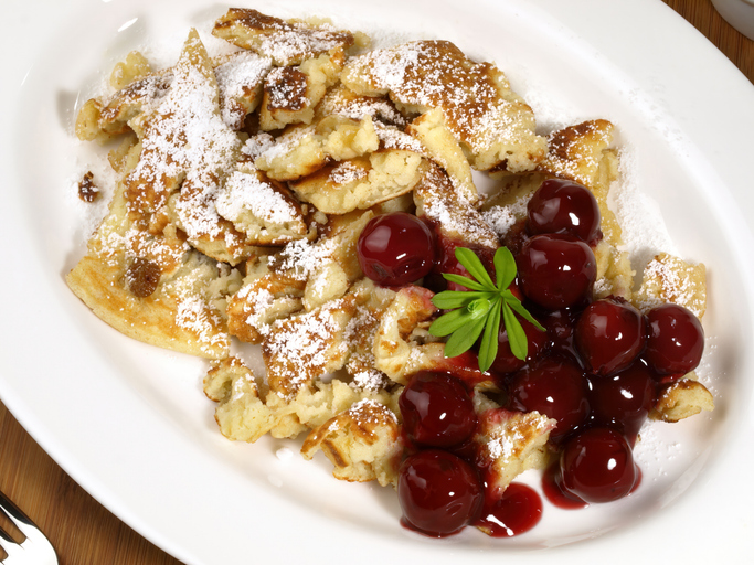 Ein weißer Teller mit Kaiserschmarrn und Rosinen, serviert mit glänzenden Sauerkirschen und garniert mit einem grünen Waldmeisterblatt.