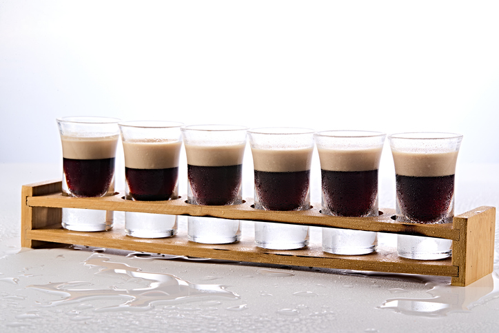 Sechs Shotgläser in einer Holzhalterung, gefüllt mit den drei charakteristischen Schichten aus Kaffeelikör, Cremelikör und Orangenlikör.