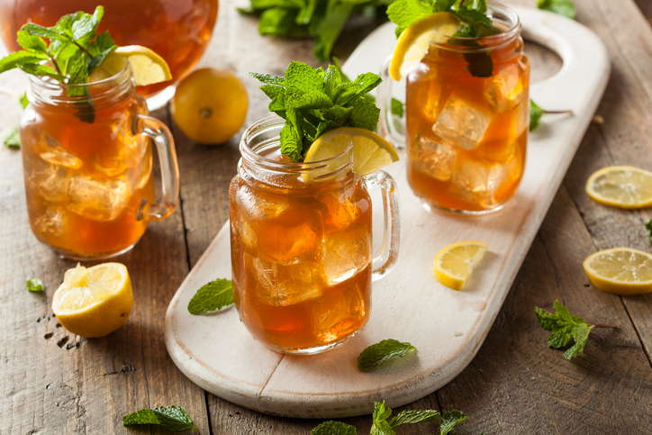 Drei Gläser mit hausgemachtem Zitroneneistee stehen mit Eiswürfeln, Zitronenstücken und Minze auf einem Brett.