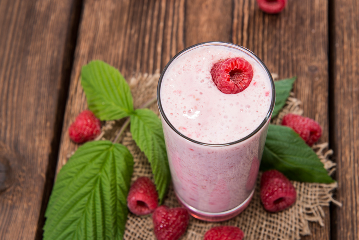 Ein hoher Himbeer-Joghurt Shake steht auf einem Holzbrett, dekoriert mit Himbeeren und grünen Blättern.