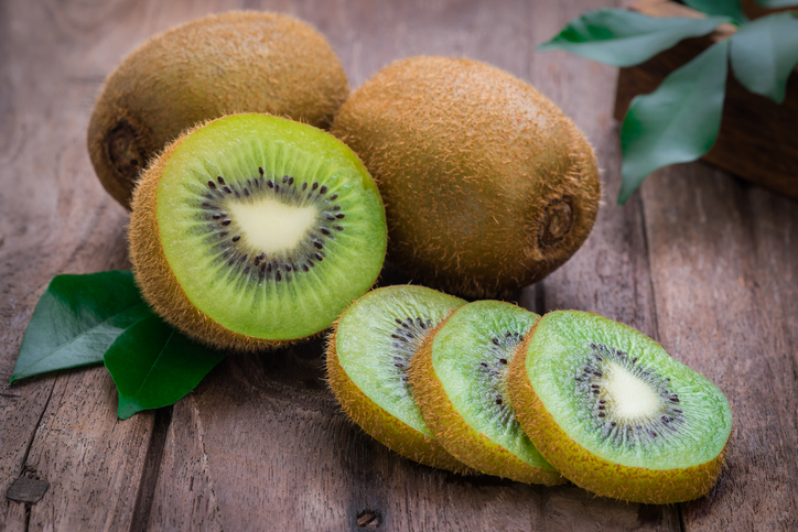 Ganze und in Scheiben geschnittene Kiwi liegen auf einem rustikalen Holztisch mit grünen Blättern daneben.