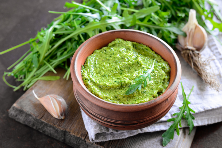 Eine Schale mit grünem Rucola-Pesto steht neben frischem Rucola und Knoblauch auf einem Holzbrett.