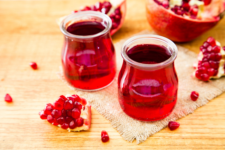 Zwei bauchige Gläser mit Granatapfel-Likör stehen auf einem Jutestoff auf hellem Holz, im Hintergrund sind ganze Granatäpfel und lose Kerne zu sehen.