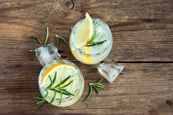 Draufsicht auf zwei Gläser Gin Tonic, dekoriert mit Zitrone und Rosmarin, umgeben von Eiswürfeln auf einem dunklen Holzuntergrund.